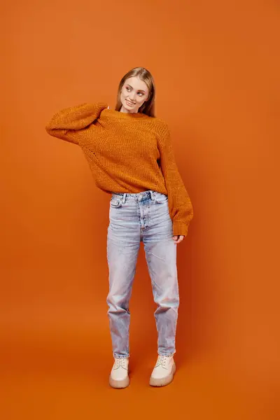 Full length of trendy blonde woman in jeans and soft knitted sweater posing on orange backdrop — Stock Photo