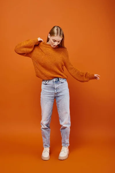 Attractive blonde woman in bright knitted sweater and jeans on orange backdrop, winter style — Stock Photo
