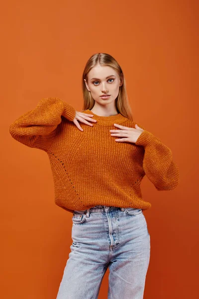 Expressive blonde woman in bright knitted sweater and blue jeans on orange backdrop, modern fashion — Stock Photo