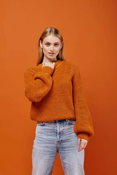 Verführerische blonde Frau in hellem Strickpullover und blauer Jeans, die vor orangefarbenem Hintergrund in die Kamera blickt — Stockfoto