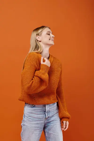 Excited blonde woman in bright sweater and blue jeans laughing and looking away on orange backdrop — Stock Photo