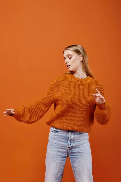 Fashionable blonde woman in bright sweater and blue jeans posing on orange backdrop, winter trend — Stock Photo
