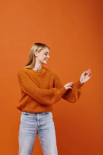 Pleased blonde woman looking at her warm and bright knitted sweater on orange backdrop, cozy winter — Stock Photo