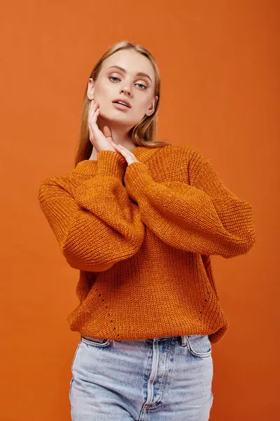 Young sensual female model in blue jeans and bright sweater looking at camera om orange backdrop — Stock Photo