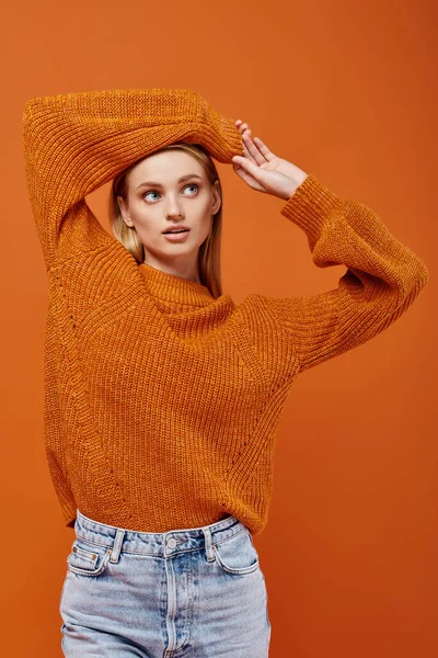 Mujer rubia joven en suéter de punto naranja posando con los brazos por encima de la cabeza en naranja, vibraciones de invierno - foto de stock