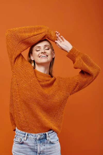 Happy blonde woman in bright winter sweater with arms above head smiling with closed eyes on orange — Stock Photo