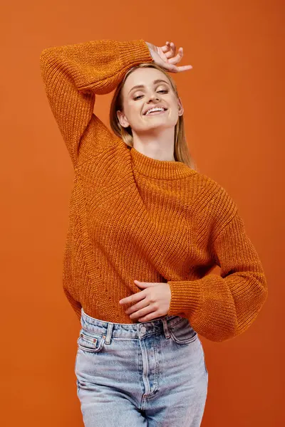 Mujer rubia feliz en suéter de invierno brillante posando con la mano por encima de la cabeza en naranja, alegría de invierno - foto de stock