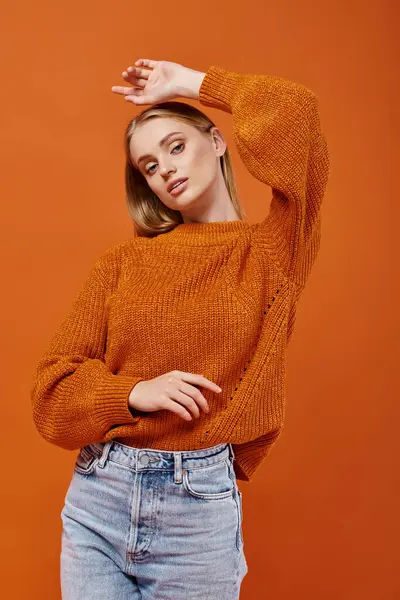 Trendy blonde woman in bright sweater posing with hand above head and looking at camera on orange — Stock Photo