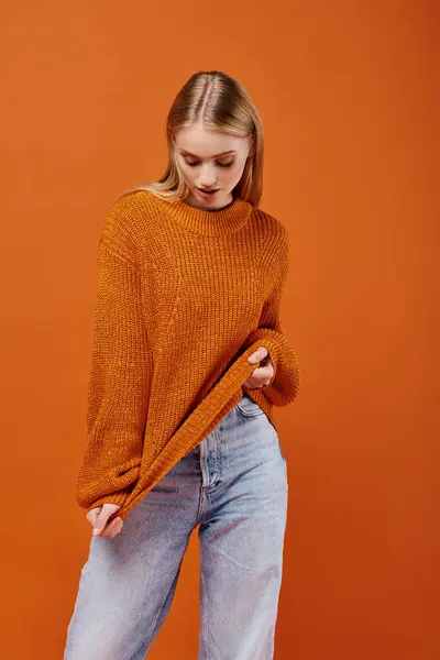 Young blonde woman in blue jeans stretching her bright warm sweater on orange backdrop, fashion — Stock Photo