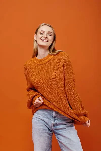 Mujer rubia alegre estirando su suéter brillante y sonriendo a la cámara en naranja, emociones de invierno - foto de stock
