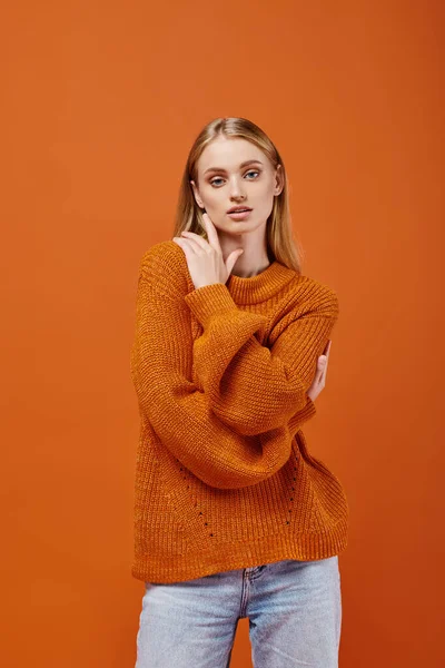 Mujer rubia pensativa en suéter de punto naranja de pie sobre fondo naranja, emociones de invierno - foto de stock