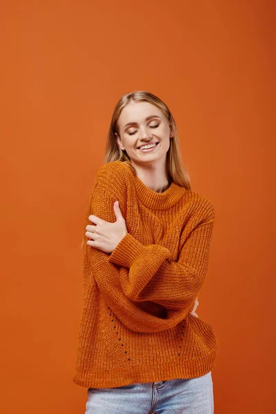 Pleased woman in warm knitted sweater hugging herself and smiling with closed eyes on orange — Stock Photo