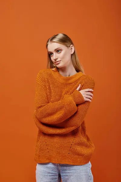 Dreamy blonde woman in bright and warm knitwear hugging herself and looking away on orange — Stock Photo
