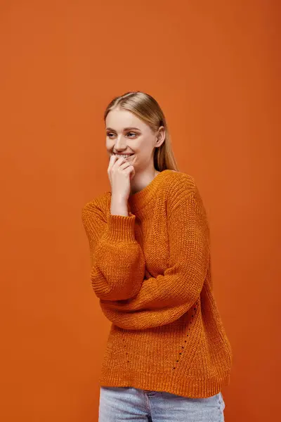 Mujer rubia sonriente en suéter de punto brillante sonriendo y mirando hacia otro lado en naranja, emociones de invierno - foto de stock