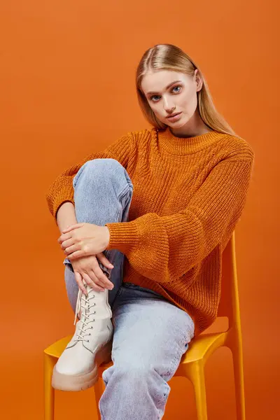 Femme blonde tendance en pull tricoté orange et jean bleu assis sur une chaise lumineuse, style hiver — Photo de stock