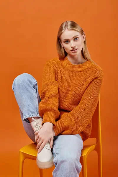 Stylish blonde woman in warm orange sweater and blue jeans sitting on bright chair, winter fashion — Stock Photo