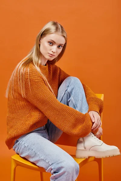 Trendige blonde Frau in orangefarbenem Strickpullover und blauer Jeans sitzt auf Stuhl und blickt in die Kamera — Stockfoto