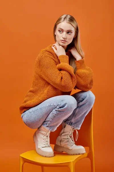 Thoughtful woman in colorful sweater and blue jeans posing on chair and looking at camera on orange — Stock Photo
