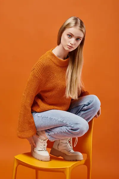 Modelo femenina rubia en suéter naranja cálido y vaqueros azules posando en la silla y mirando a la cámara - foto de stock