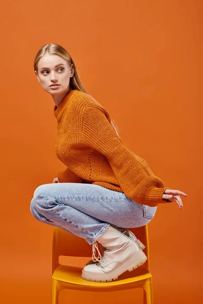Atractiva mujer en suéter de punto de invierno y jeans posando en la silla mientras mira hacia otro lado en naranja - foto de stock