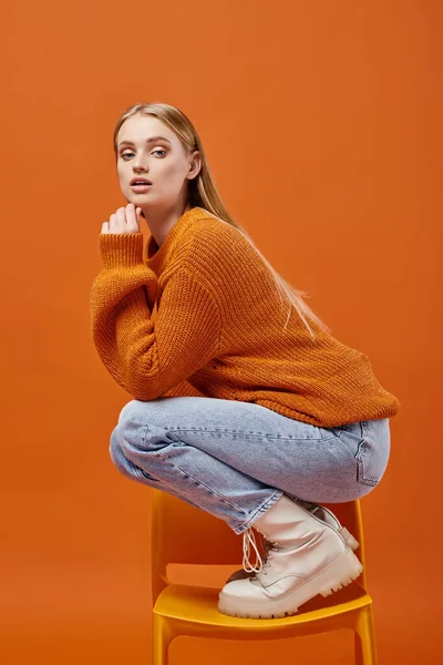 Expressive woman in knitted winter sweater and jeans posing on chair and looking at camera on orange — Stock Photo
