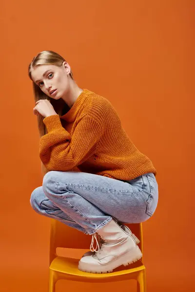 Jeune femme blonde en pull d'hiver chaud et jeans posant sur la chaise et regardant la caméra sur orange — Photo de stock