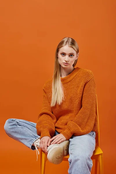 Mujer rubia de moda en suéter de invierno caliente y vaqueros sentados en la silla mirando a la cámara en naranja - foto de stock