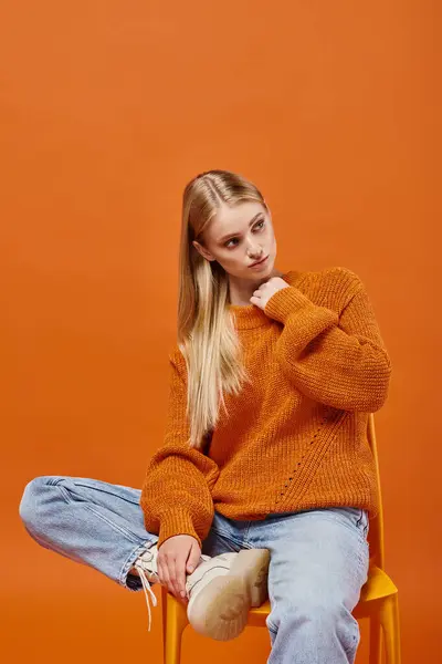 Mujer rubia pensativa en suéter de invierno brillante y jeans sentados en la silla y mirando hacia otro lado en naranja - foto de stock