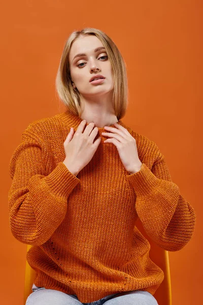 Выразительная блондинка в ярком зимнем свитере сидит на стуле, глядя на камеру на оранжевом — стоковое фото