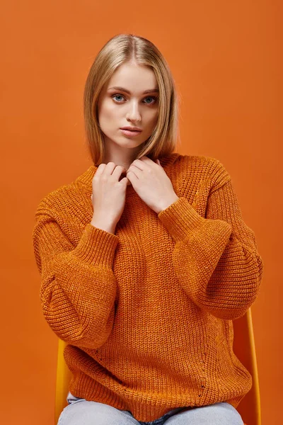 Joven mujer rubia en suéter naranja sentado en la silla mientras mira a la cámara en el fondo brillante - foto de stock