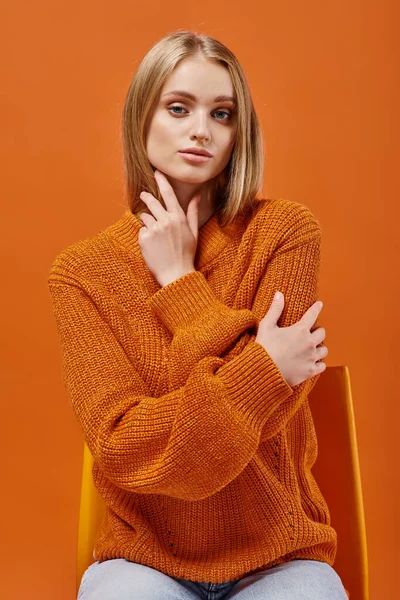 Fashionable blonde woman in orange sweater sitting on chair and looking at camera on bright backdrop — Stock Photo