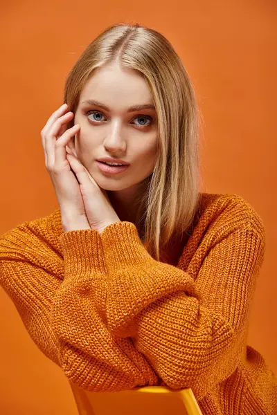 Задумчивая блондинка в ярком уютном трикотажном свитере с натуральным макияжем, смотрящая в камеру на оранжевый — стоковое фото