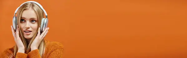 Inspirierte Frau in warmem Strickpullover, die Musik über Kopfhörer auf orangefarbenem, waagerechtem Banner hört — Stockfoto