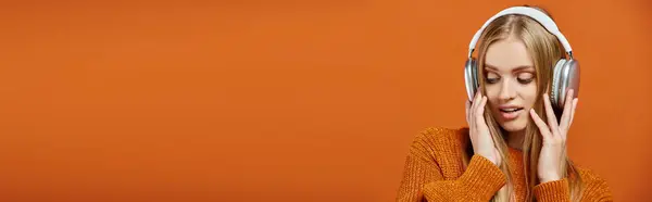 Блондинка в оранжевом трикотаже свитер и наушники слушать музыку на ярком фоне, баннер — стоковое фото