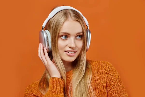 Positive blonde woman in bright winter sweater and headphones listening music on orange backdrop — Stock Photo