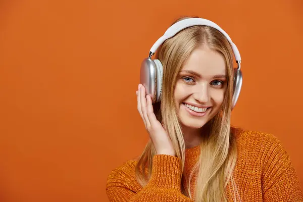 Femme blonde gaie en pull d'hiver orange et écouteurs écoutant de la musique et regardant la caméra — Photo de stock