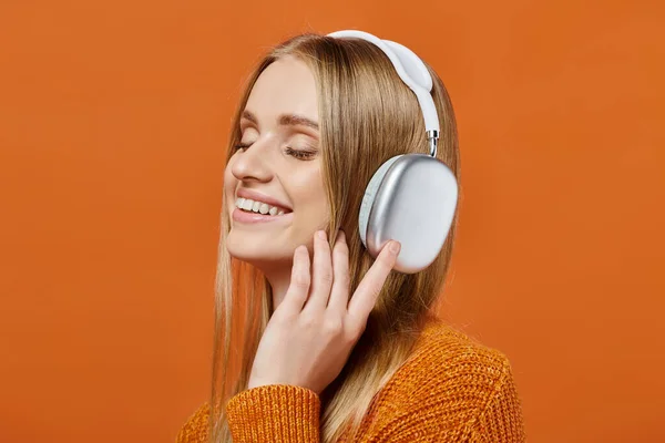 Mujer rubia encantada en suéter brillante y auriculares escuchando música con los ojos cerrados en naranja - foto de stock