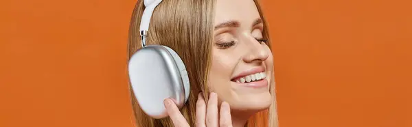 Портрет довольной блондинки в прослушивании музыки в наушниках с закрытыми глазами на оранжевый, баннер — стоковое фото