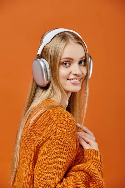 Jeune femme joyeuse en pull d'hiver orange et écouteurs écoutant de la musique et souriant à la caméra — Photo de stock