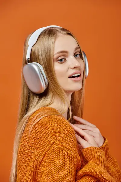 Amazed blonde woman in bright sweater and headphones listening music and looking away on orange — Stock Photo