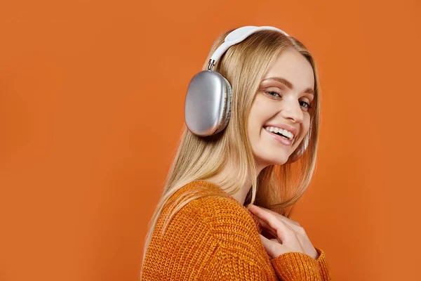 Femme blonde gaie en chandail coloré et écouteurs écoutant de la musique sur orange, vibes d'hiver — Photo de stock
