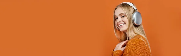 Joyeuse femme blonde en pull coloré et écouteurs écoutant de la musique sur orange, bannière horizontale — Photo de stock