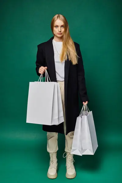 Full length of trendy blonde woman in black coat with white shopping bags on green, seasonal sales — Stock Photo