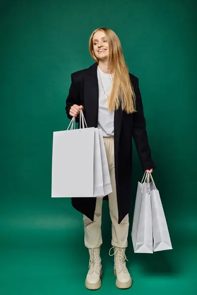 Longitud completa de mujer rubia alegre en abrigo negro con bolsas de compras blancas en verde, las ventas de invierno - foto de stock