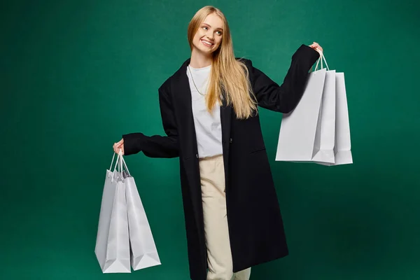 Smiling blonde woman in black trendy coat holding white shopping bags on green, winter joy — Stock Photo