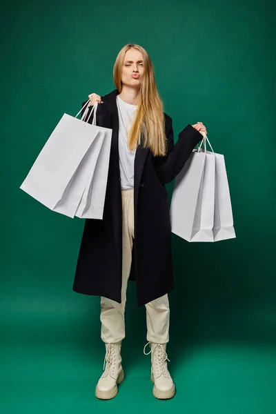 Trendy blonde woman in black coat holding white shopping bags and pouting lips on green, full length — Stock Photo