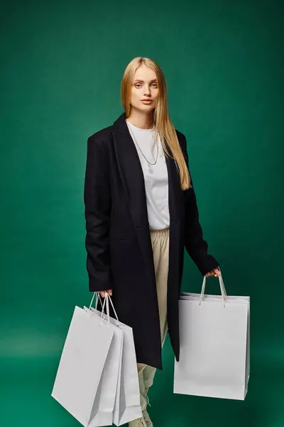 Confident blonde woman in black coat holding white shopping bags and looking at camera on green — Stock Photo