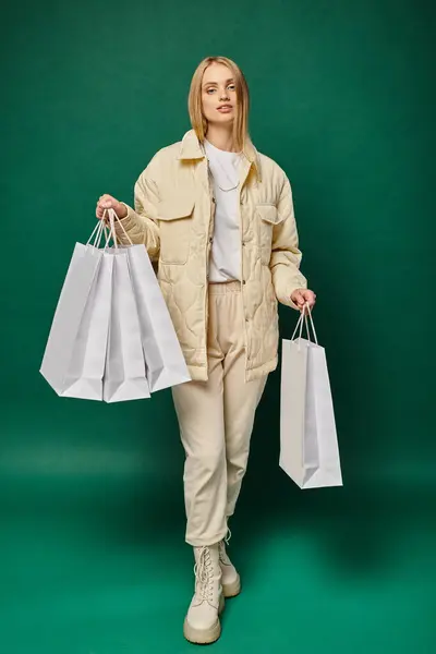 Full length of blonde woman in trendy winter clothes holding white shopping bags on green backdrop — Stock Photo