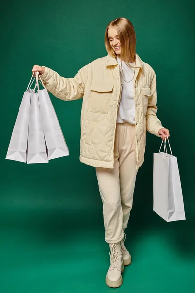 Longitud completa de mujer rubia alegre en traje de invierno de moda sosteniendo bolsas de compras blancas en verde - foto de stock