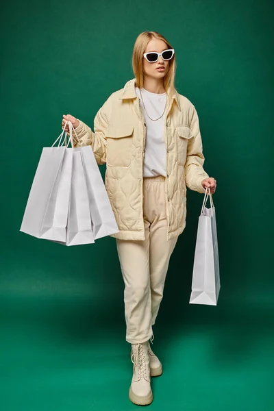 Elegante donna bionda in inverno indossare e occhiali da sole a piedi con borse della spesa su verde, a tutta lunghezza — Foto stock
